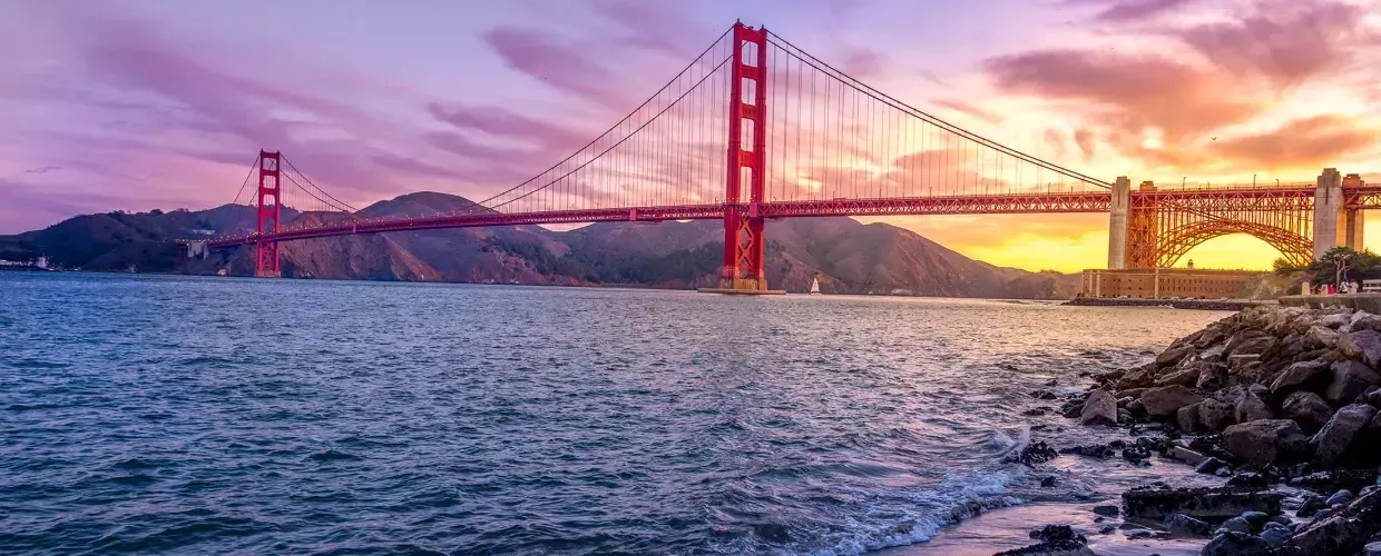 A Ponte Golden Gate ao pôr do sol 和 um céu multicolorido e a Baía de São Francisco em primeiro plano.