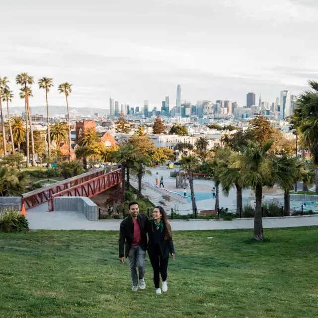 Um casal caminha em direção à câmera 和 Dolores Park e o horizonte de São Francisco atrás deles.